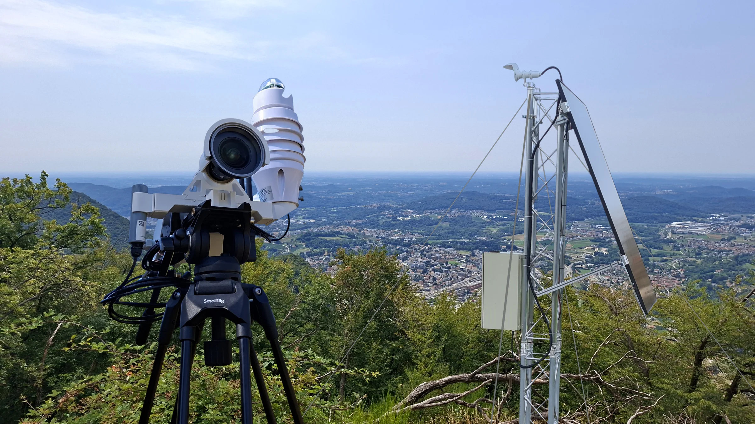 HUVI.NOMAD su un treppiede in primo piano, dietro una torre sensore HUVI.GUARDIAN con telecamera e pannello solare, sullo sfondo il Ticino
